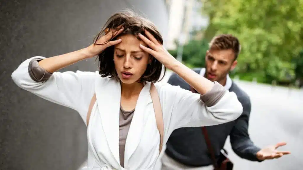 Stressed woman holding her head while a man argues behind her outdoors.