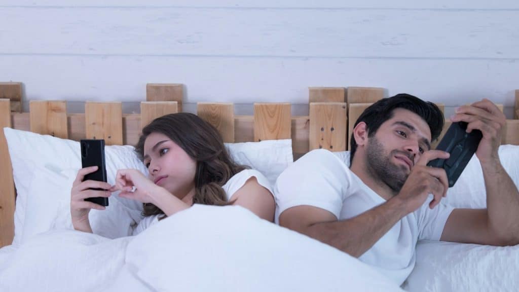 Couple lying in bed back-to-back, both preoccupied with their smartphones.