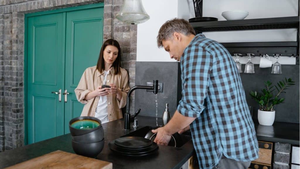 Man washing dishes at a kitchen sink while a woman stands nearby.