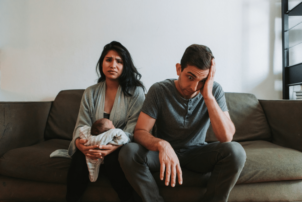 A young, stressed-looking couple sits on a couch, with the woman holding a sleeping newborn baby and the man holding his head.
