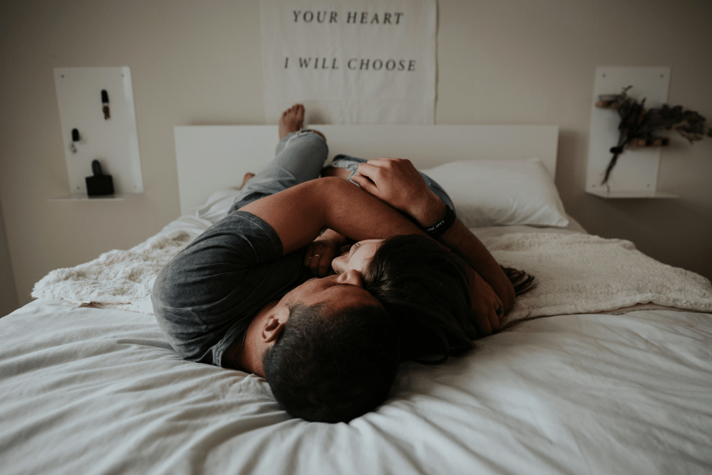 A couple lying in bed, embracing, with the man's head on the woman's chest and a poster on the wall.