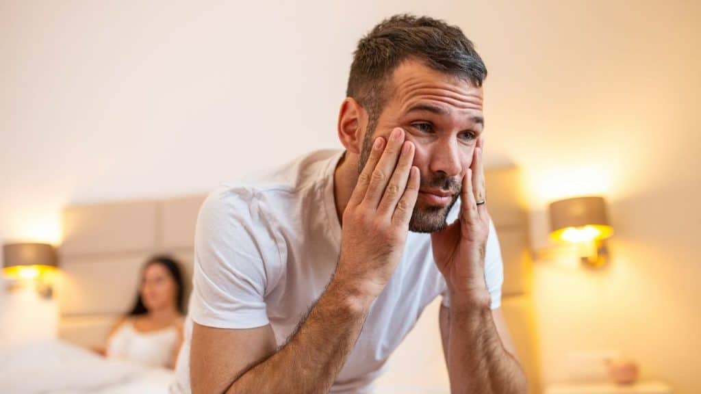 A tired man rubs his face in a bedroom with a woman sitting in the background.