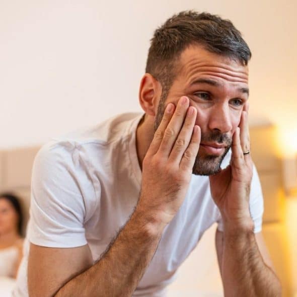 A tired man rubs his face in a bedroom with a woman sitting in the background.