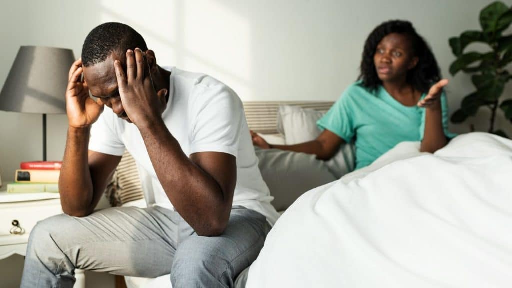 A distressed man sits on a bed while a frustrated woman gestures in the background.