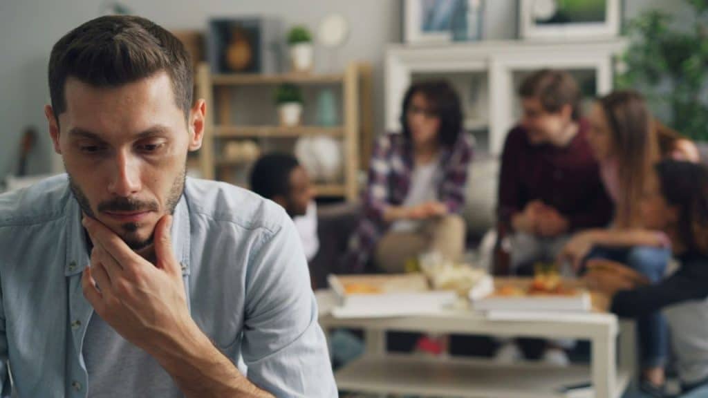 A pensive man holds his chin while a group of people socializes behind him.