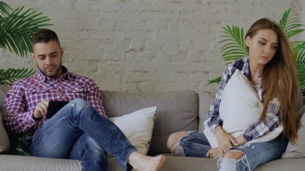 A couple sits separately on a couch; the man uses a tablet and the woman looks away.