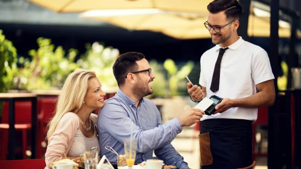 A smiling man hands a credit card to a waiter while a woman watches happily at a cafe.