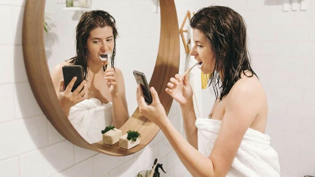 A woman in a towel brushes her teeth and looks at her phone in a mirror.