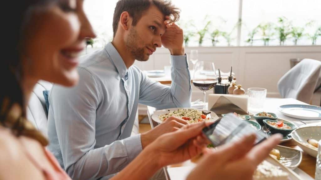 A man looks frustrated at a dining table while a woman smiles, looking at her phone.