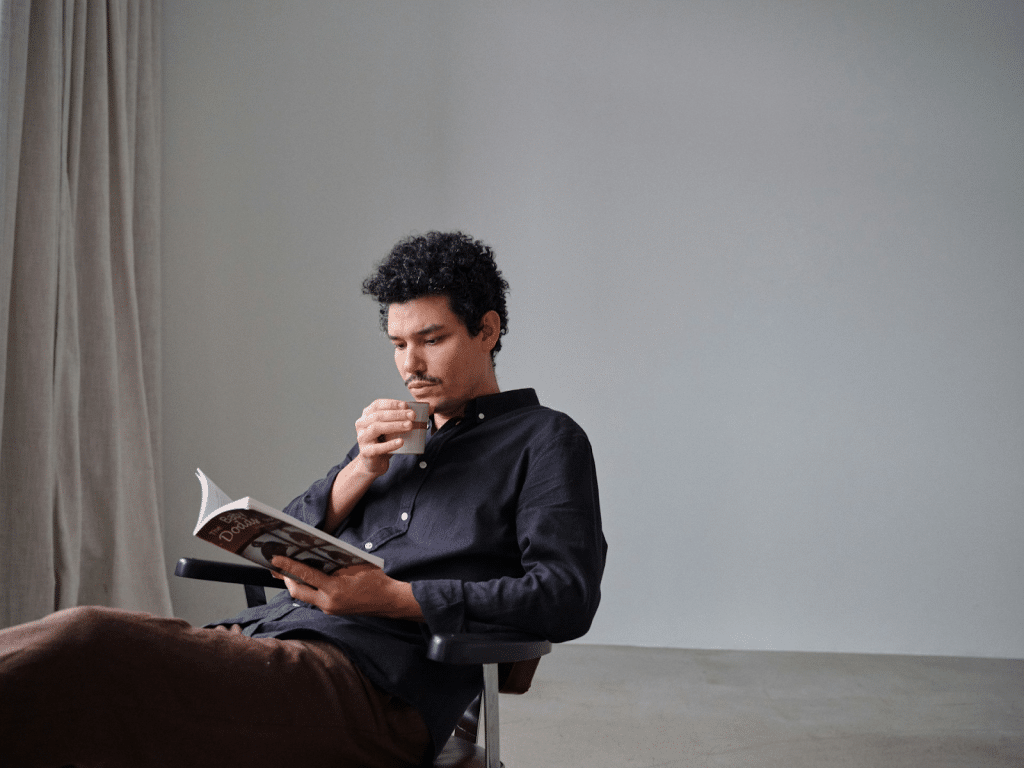 A man with curly hair and a mustache sitting in a chair, reading a book and drinking from a small cup.
