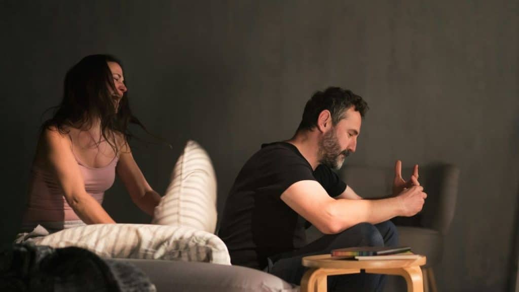 A distressed man sits looking down while a woman throws a pillow in the background.