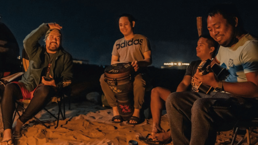 Four men sitting on a sandy beach at night, playing music around a campfire.