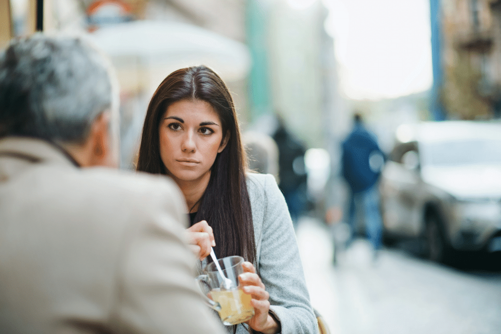A woman with long, dark hair listening intently to a person across the table while stirring a drink at an outdoor cafe.