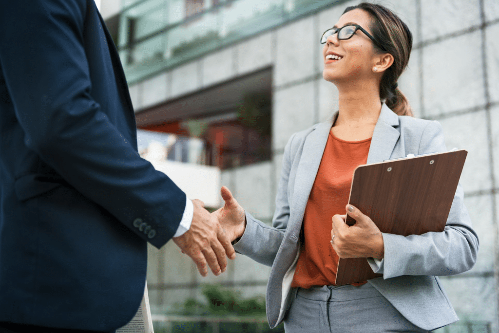 A professional woman in a grey suit and glasses smiling while shaking hands with a man outside a modern building.
