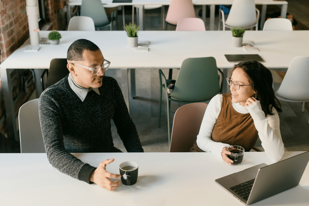 Two people wearing glasses are sitting at a white table, talking and drinking coffee with a laptop present in a modern, open-office setting.
