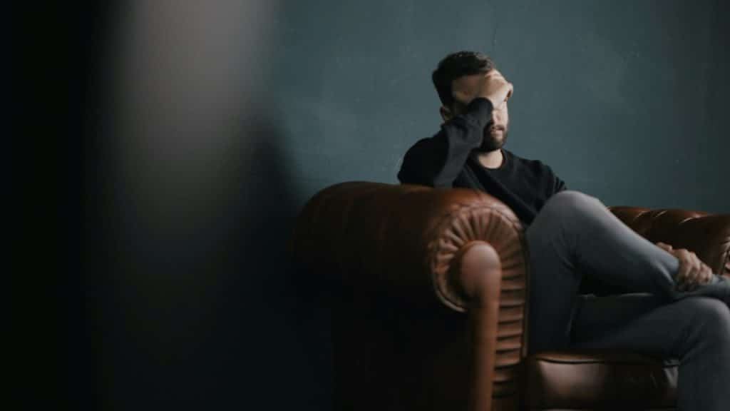 A man holds his head while sitting on a sofa.