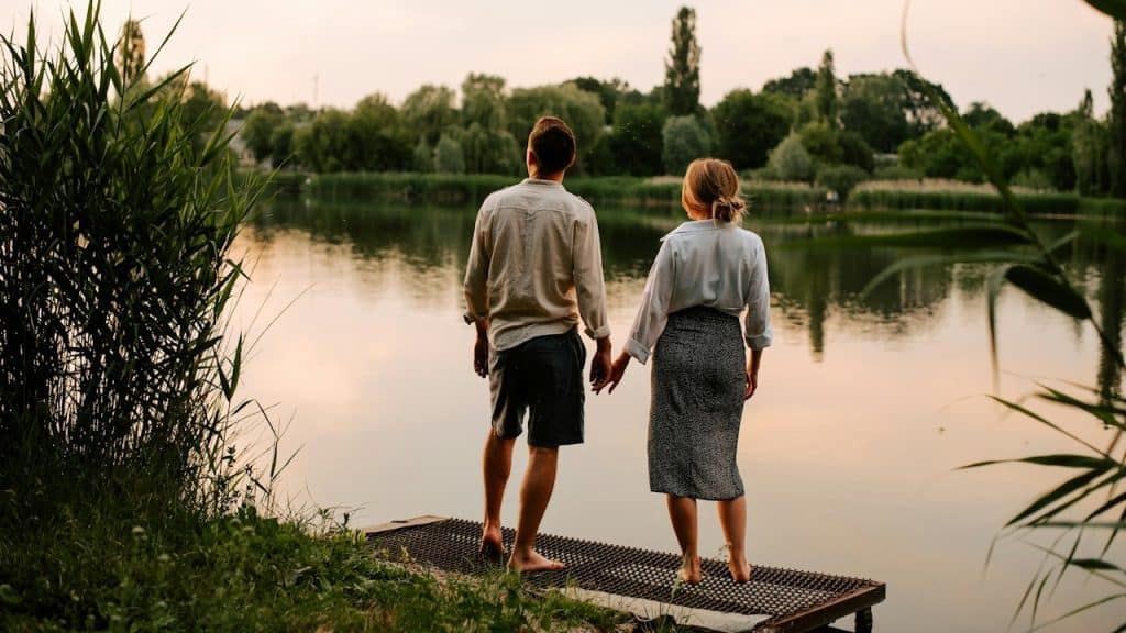 A couple holding hands and standing on a deck over a lake.