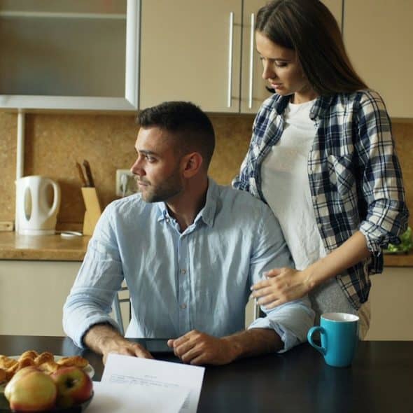Woman standing and gently touching the shoulder of a man who is sitting at a kitchen table looking upset.