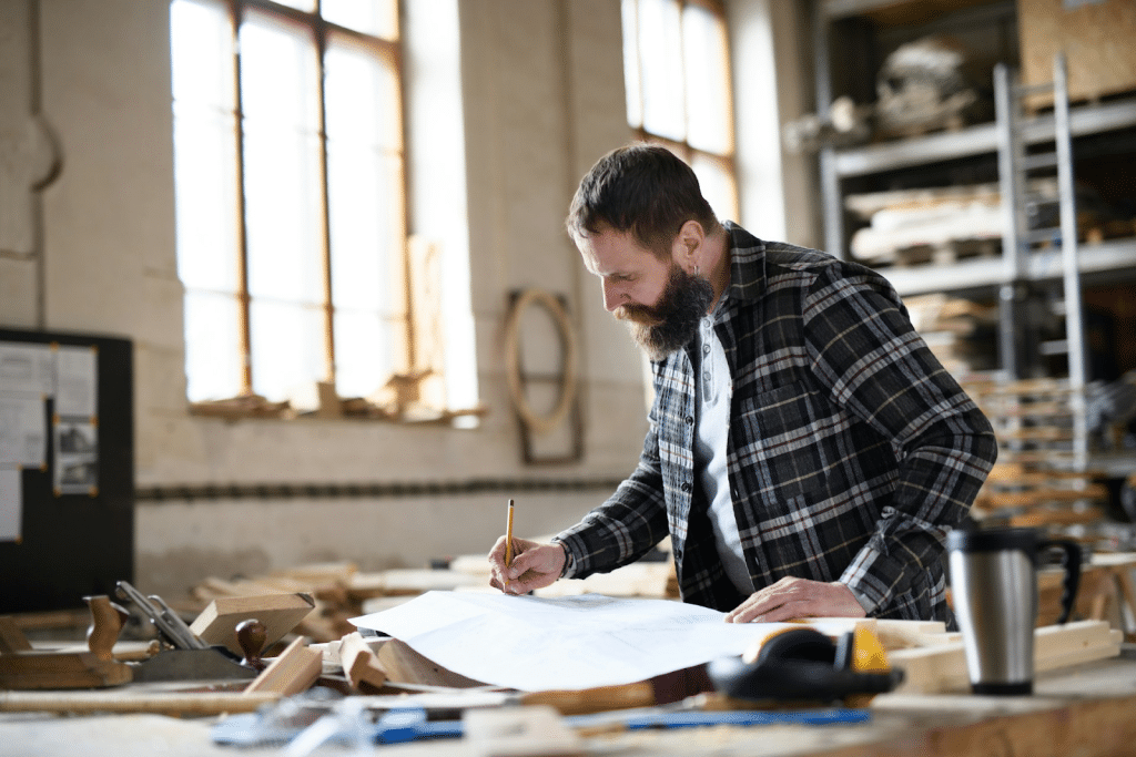 Bearded carpenter in a plaid shirt reviewing blueprints on a workbench in a bright workshop.