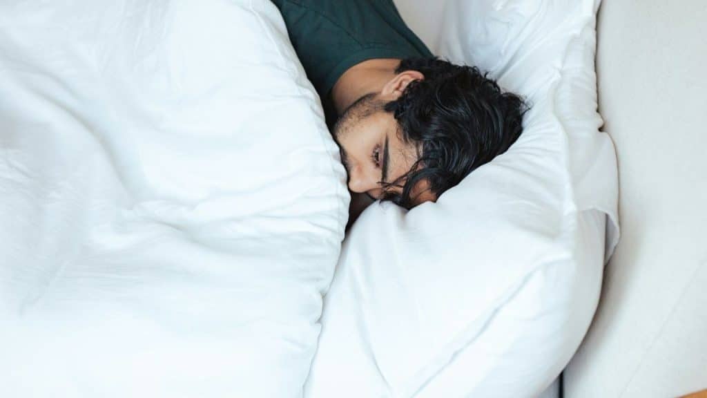 A man with dark hair is partially buried under a white comforter in bed.
