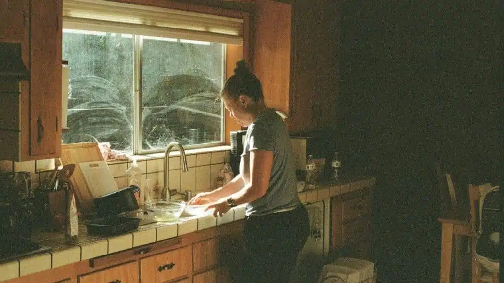 A woman washing dishes in a sunlit kitchen sink with a window overlooking greenery.