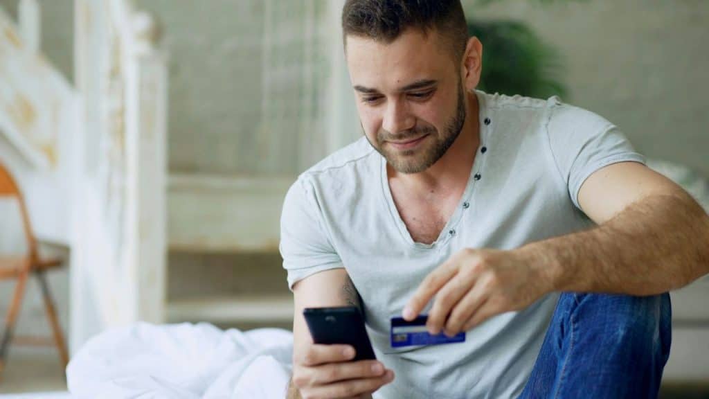 A smiling bearded man in a white shirt holds a credit card while looking at a phone.