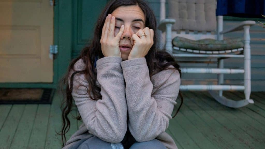 A woman sits on a porch, covering her eyes and face with both hands.