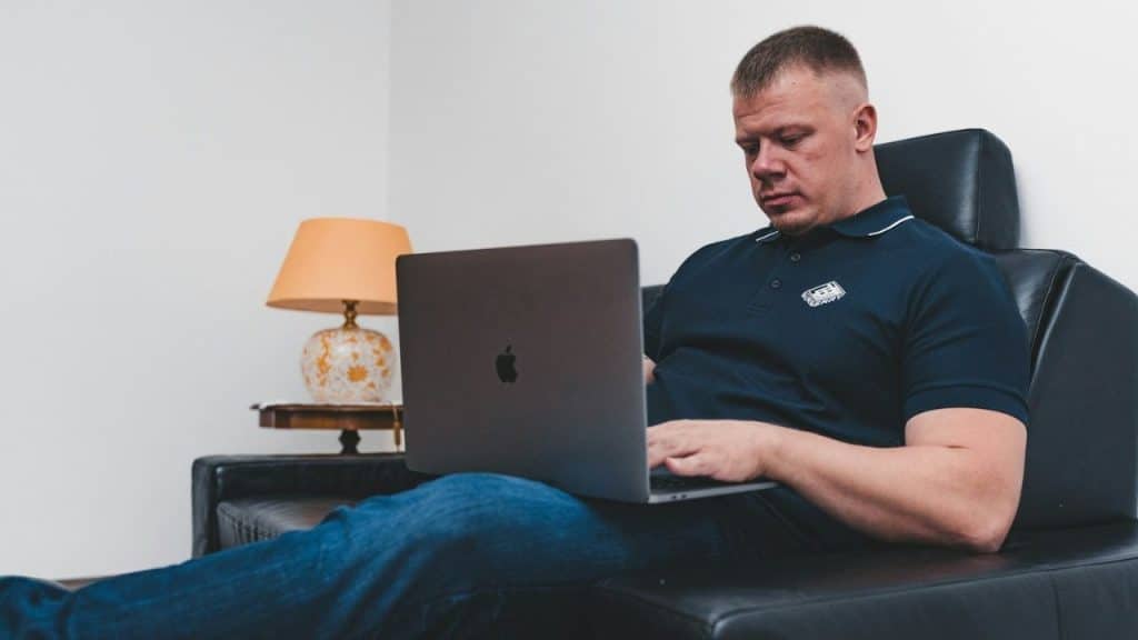 A man in a polo shirt sits on a black couch, working intently on a laptop.