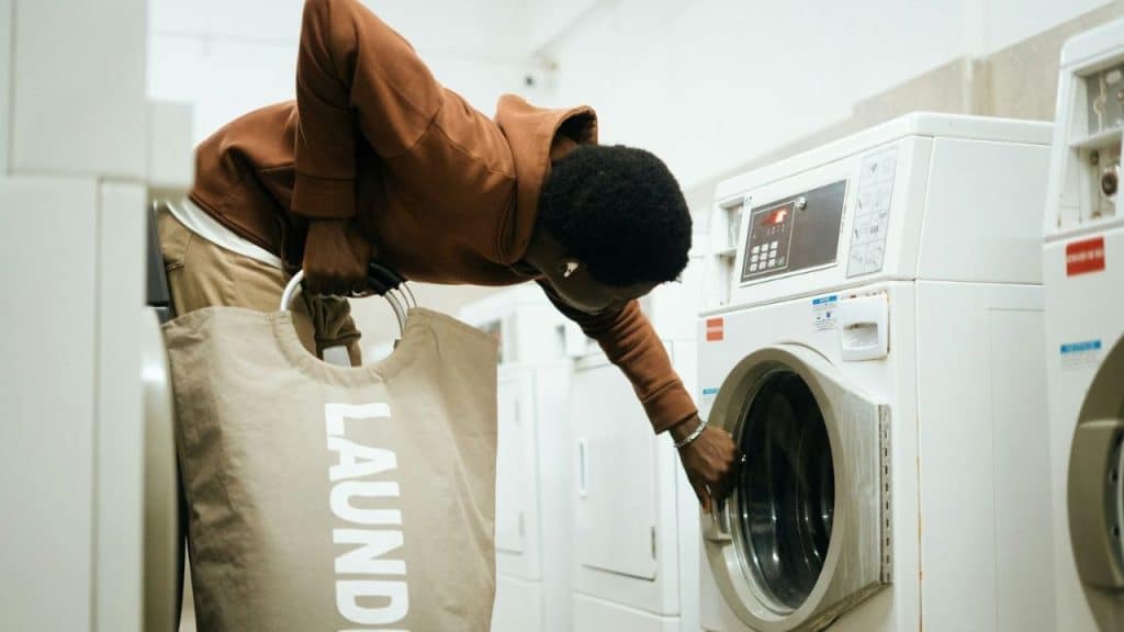 A person in a brown hoodie opens a washing machine while holding a large "LAUNDRY" bag.