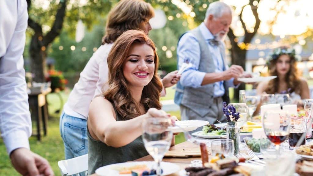 A smiling woman with long brown hair reaches for food at an outdoor dinner table.
