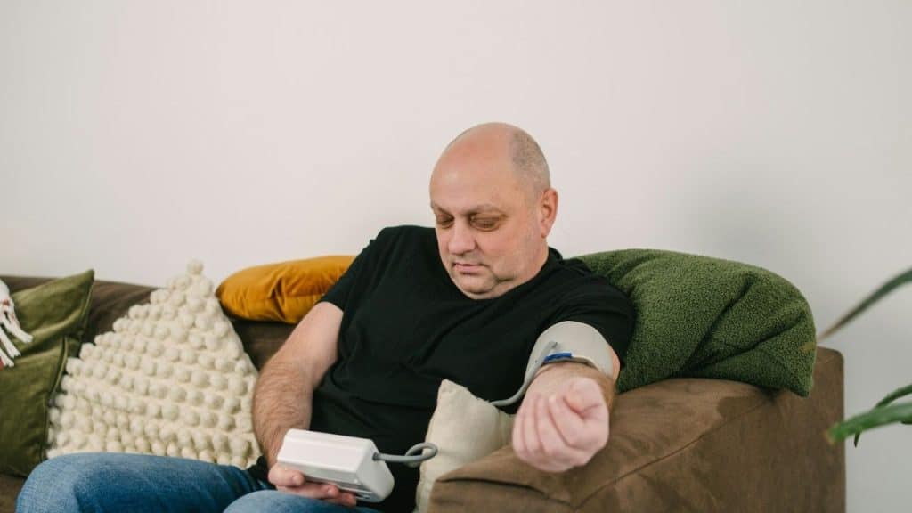 A bald man sits on a brown couch, measuring his blood pressure with a home monitor.