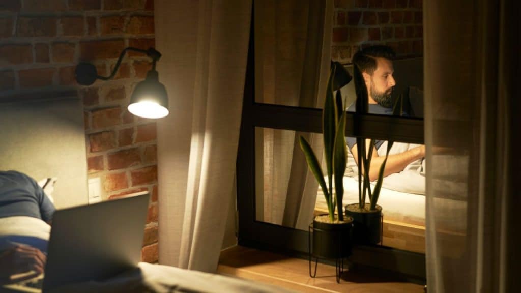 Bearded man sits in bed at night, reflected in a window next to a brightly lit brick wall.