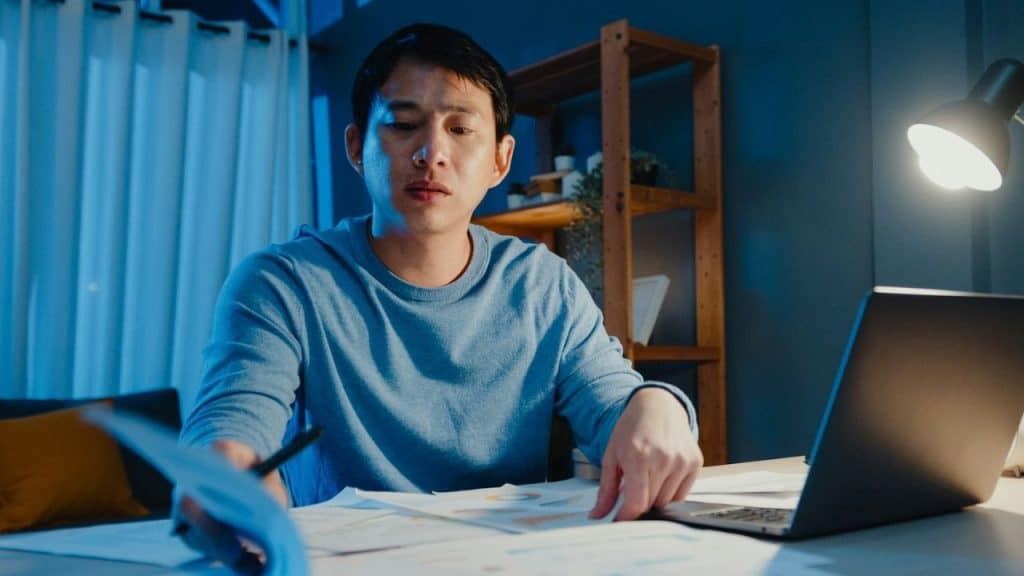 Man in a blue shirt works at a desk, looking at papers under a bright desk lamp.