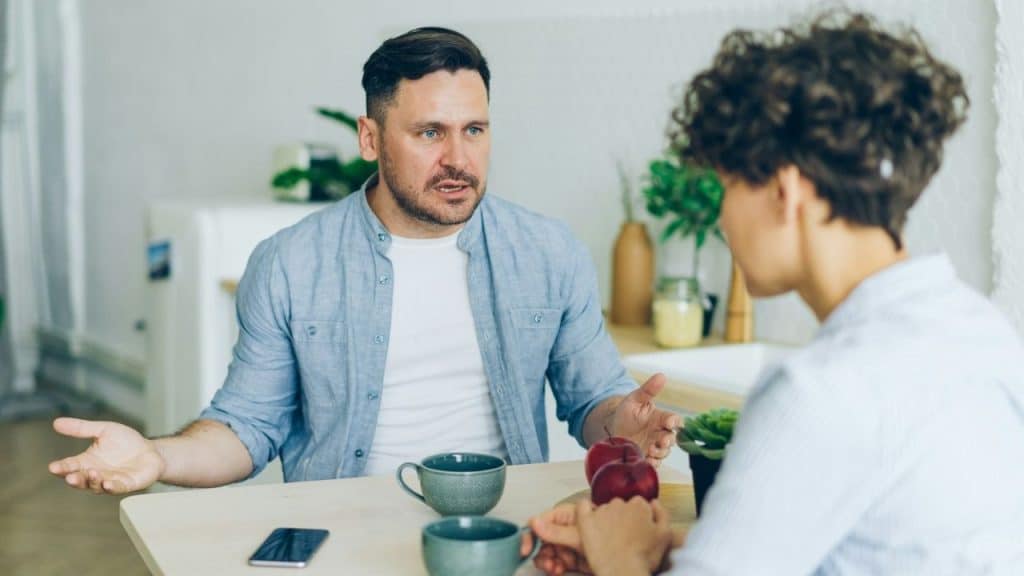 Man in a blue shirt talking heatedly across a table to a woman seen from behind.