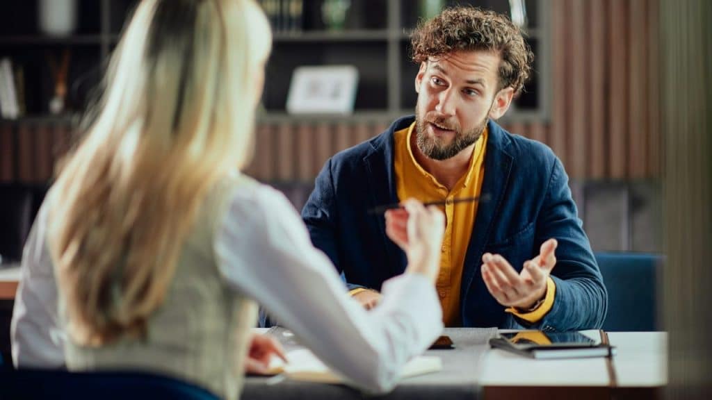 Bearded man in a navy jacket and yellow shirt speaks to a woman across a table.