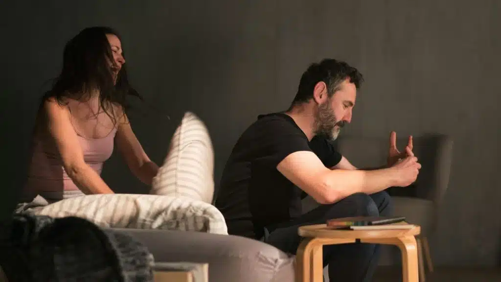 Woman raises a pillow as if to hit a seated man who is looking away with clasped hands.