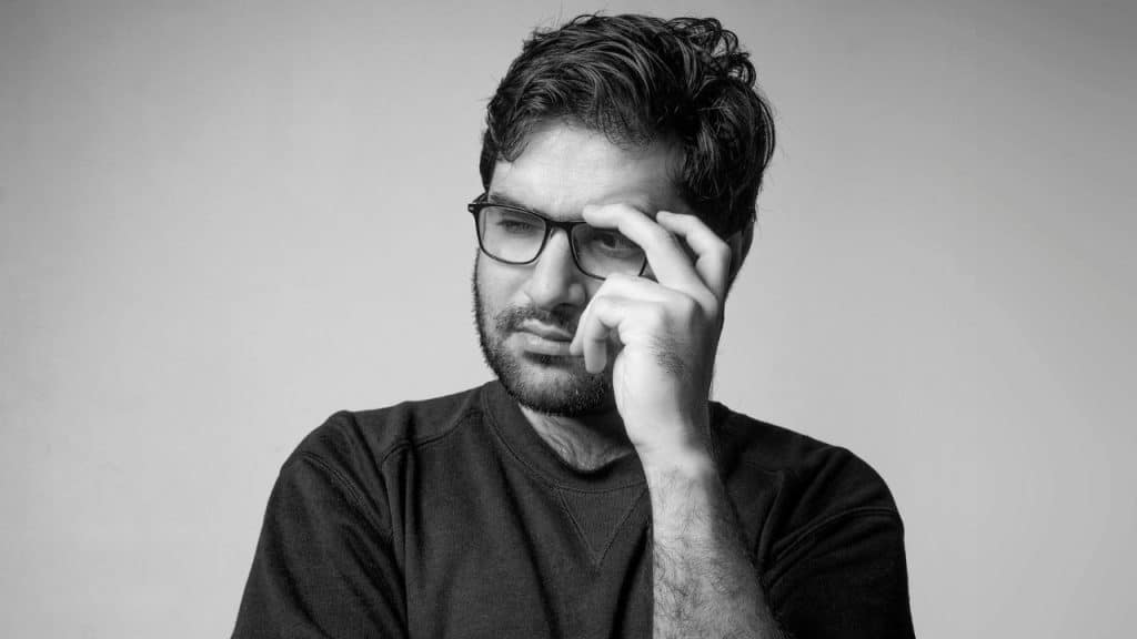 Black and white photo of a bearded man in glasses touching his forehead thoughtfully.