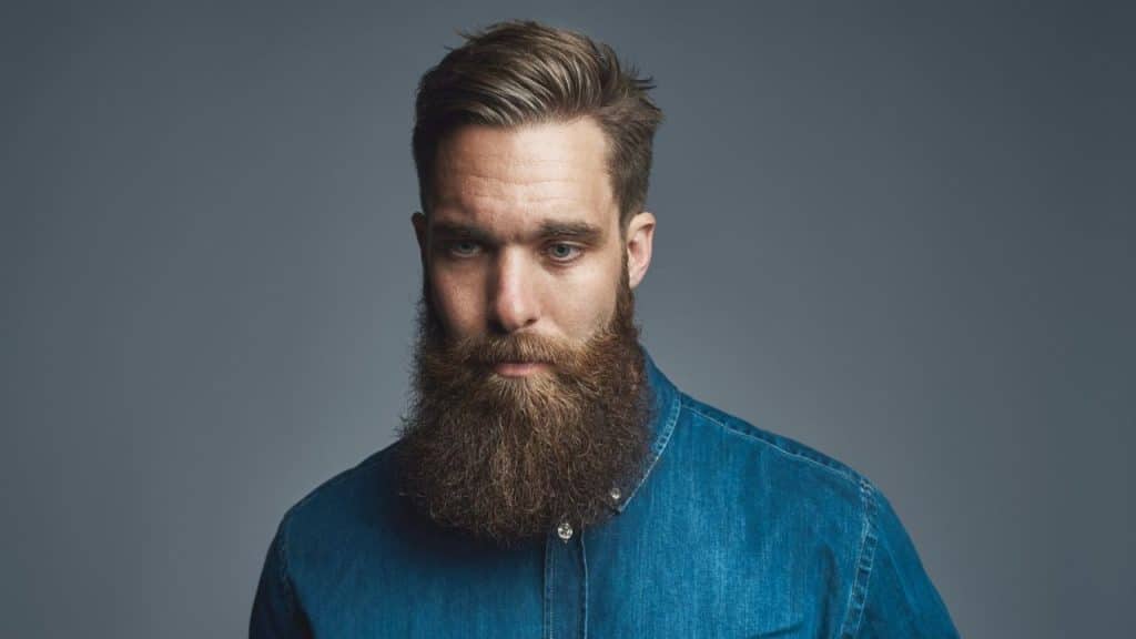 A man with a full, long brown beard and a denim shirt looks downward.