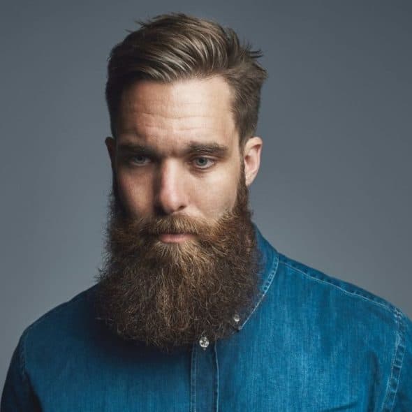 A man with a full, long brown beard and a denim shirt looks downward.