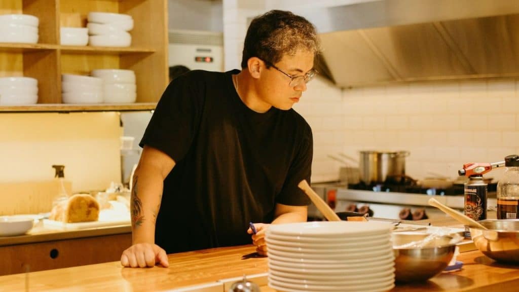 A man wearing glasses and a black shirt leans on a counter in a commercial kitchen.