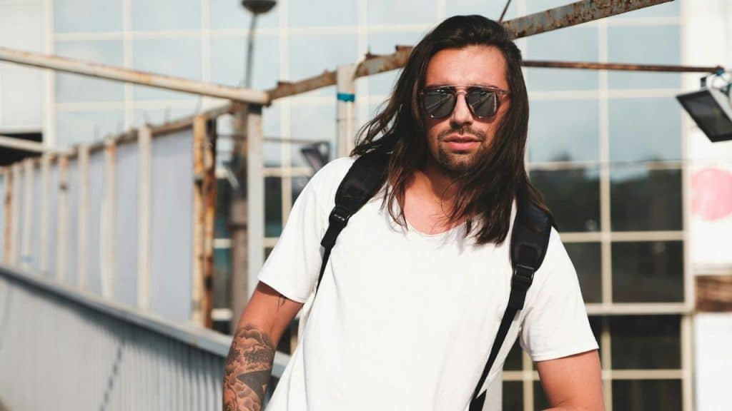 A man with long dark hair, sunglasses, and a backpack stands outdoors, looking forward.