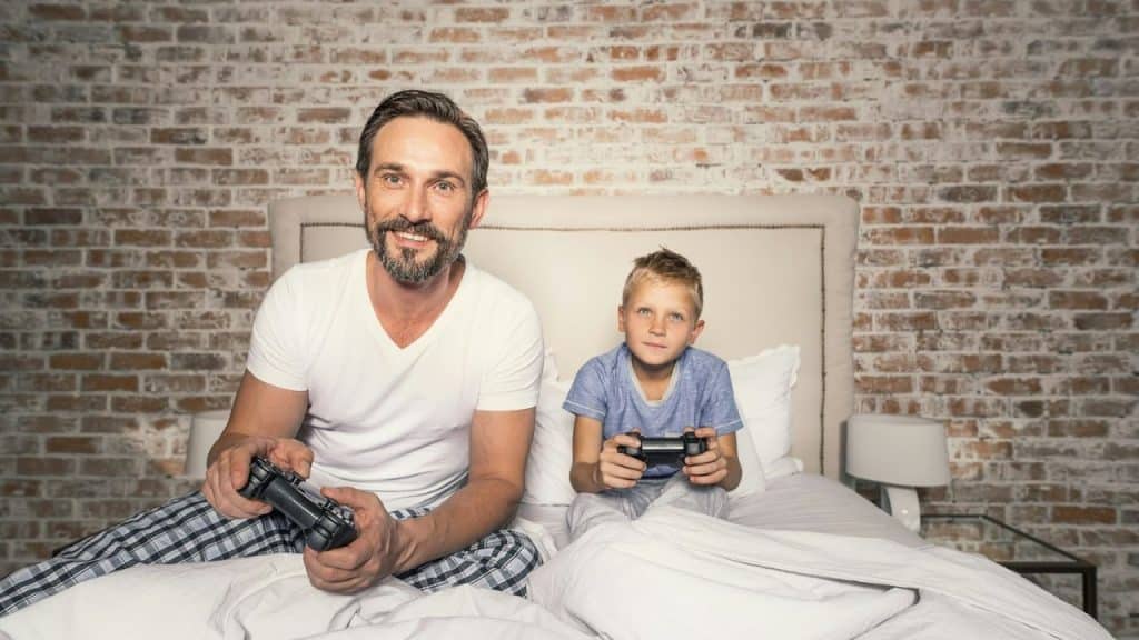 A smiling father and son sit on a bed with a brick wall background, holding video game controllers.