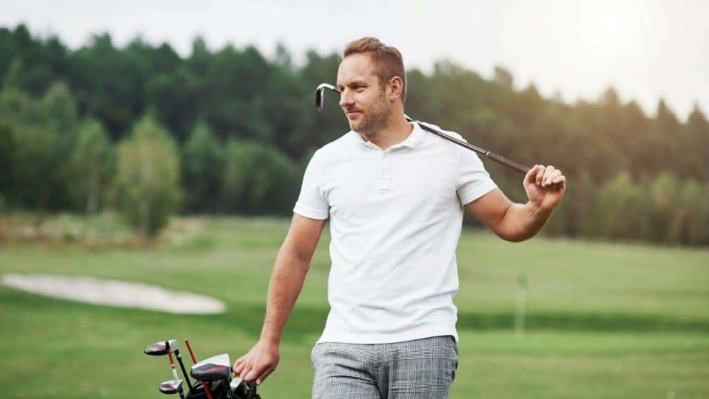 A man in a white polo shirt carries a golf club and bag on a grassy course.
