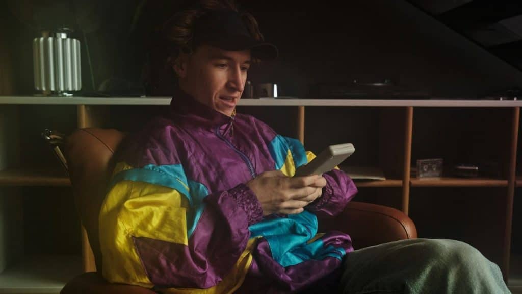 A person in a colorful 90s-style jacket and a black cap plays a handheld video game.