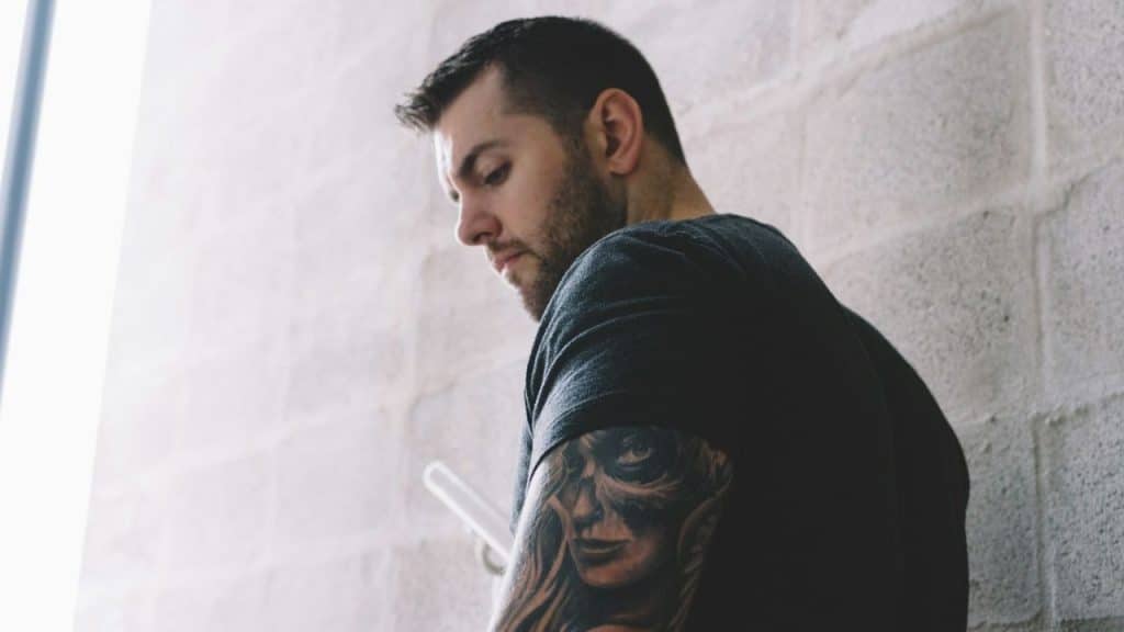 A man with short dark hair and a detailed arm tattoo looks down against a light wall.