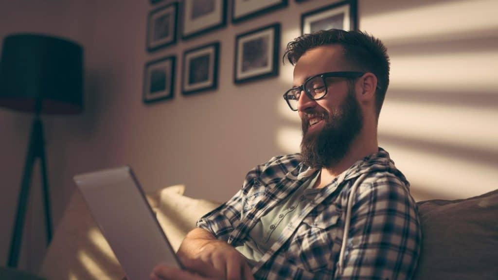 A bearded man in glasses smiles while looking at a tablet on a couch indoors.