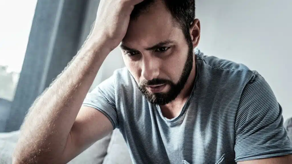 A close-up of a worried or distressed bearded man holding his head with one hand.
