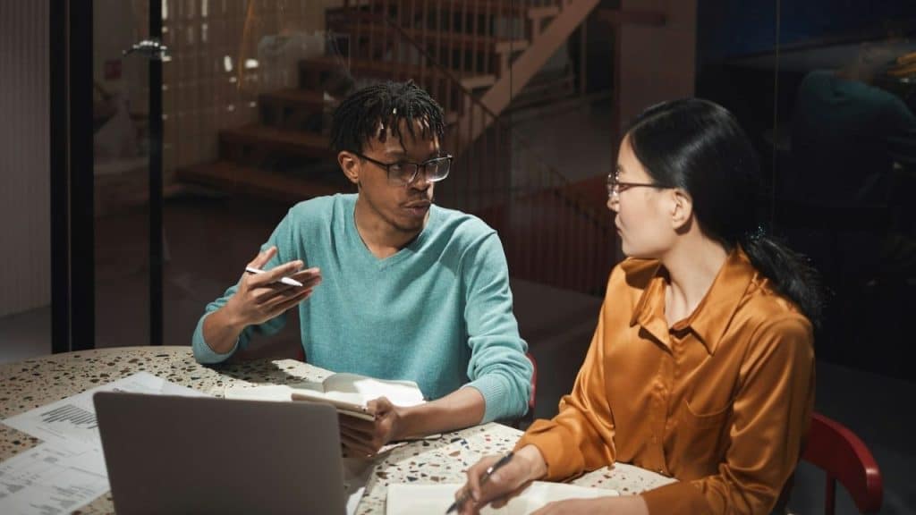 A man speaks while holding a notepad as a woman writes, sitting at a table together.