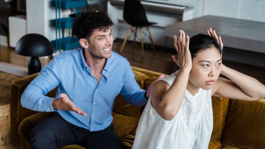 A man argues aggressively next to a woman who is covering her ears and looking upset.