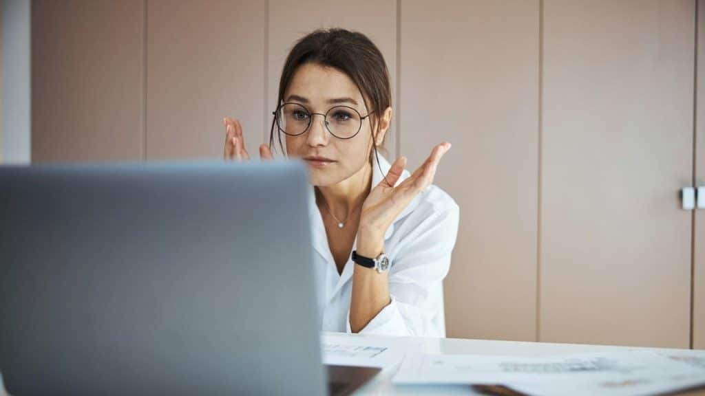 A woman with glasses looks confused at a laptop screen, gesturing with both hands.